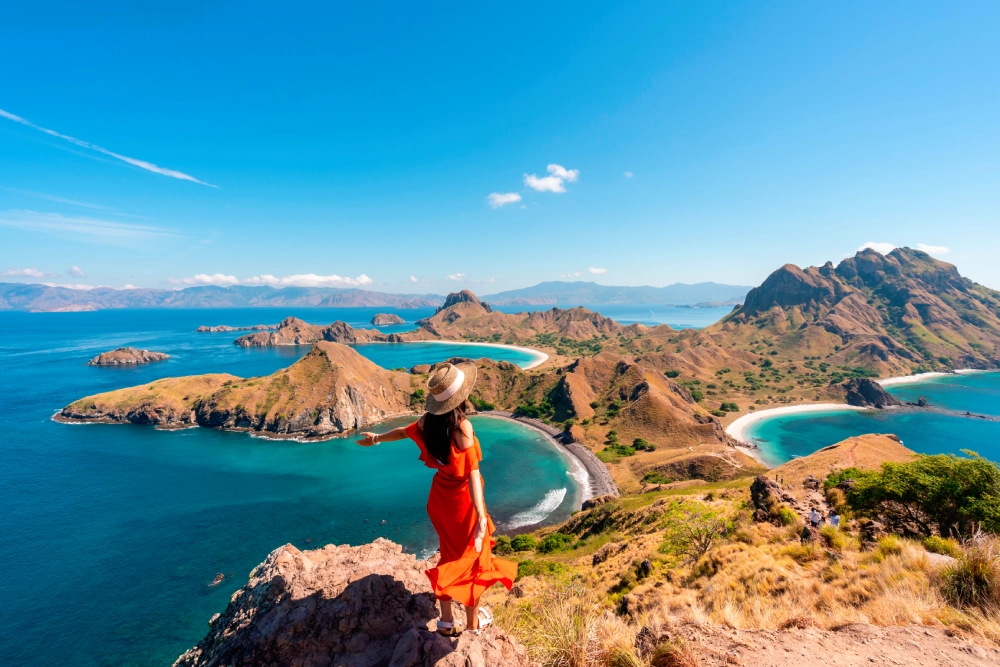 A person in a red dress and sun hat stands on a cliff overlooking a coastal landscape with blue sea and small islands.