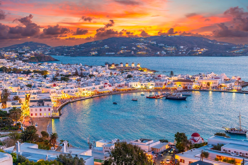 Scenic view of Mykonos town at sunset, featuring a vibrant harbor, white buildings, and the Aegean Sea under a colorful sky.