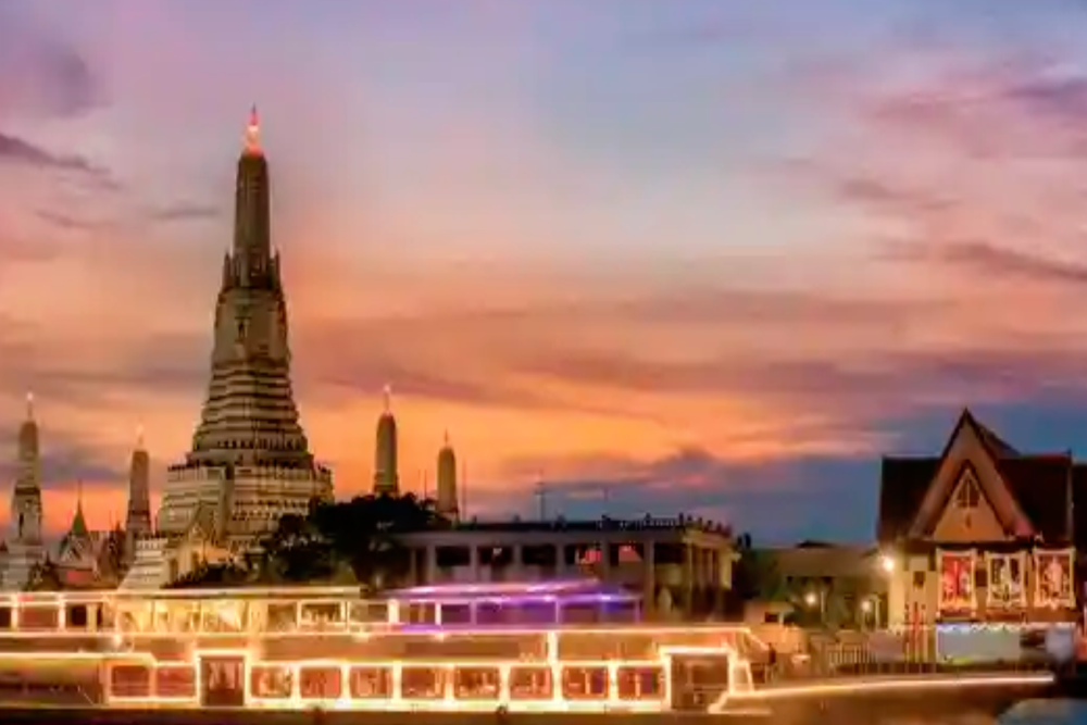 Wat Arun temple at sunset with a brightly lit boat passing on the Chao Phraya River, under a colorful sky.