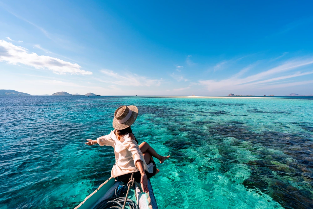 Person in a hat sitting on a boat's bow, pointing toward the clear turquoise sea with distant islands under a bright blue sky.