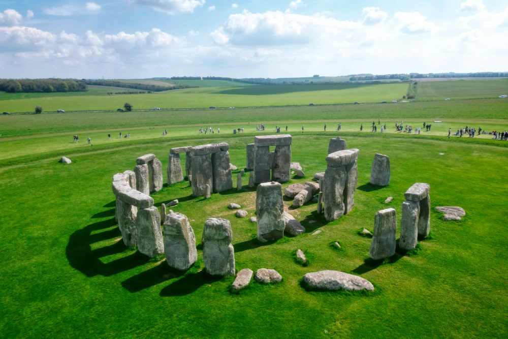 Aerial view of Stonehenge, showing a circular arrangement of large standing stones on a grassy field with scattered visitors and distant green landscape.