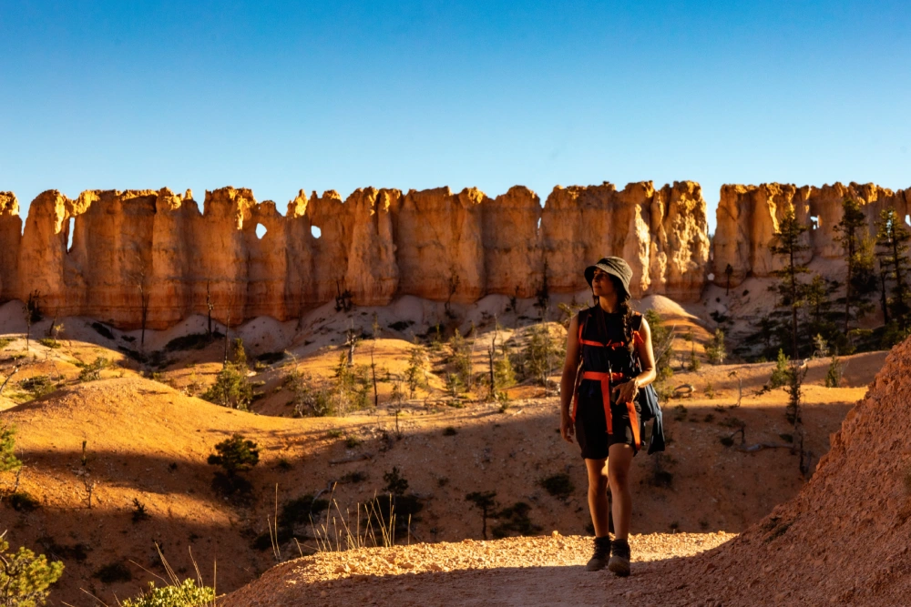 Hiker in backpack and hat walks along a trail with sunlit orange rock formations in the background, under a clear blue sky.