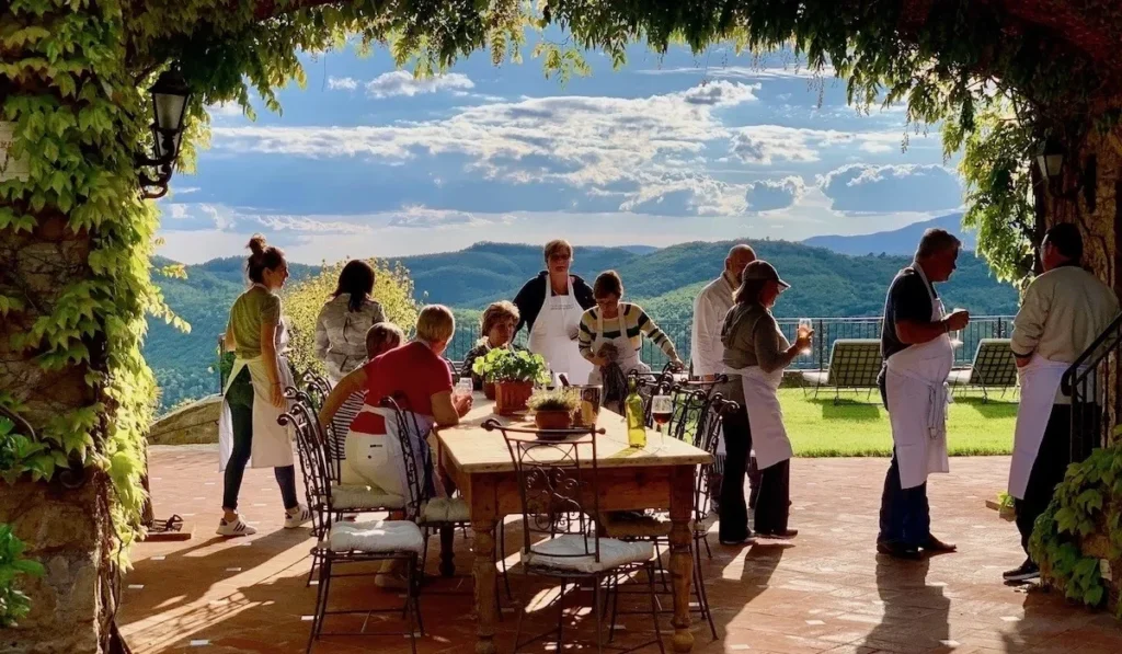A group of people, some wearing aprons, gather around a wooden table under a vine-covered archway, with a scenic mountain view in the background.